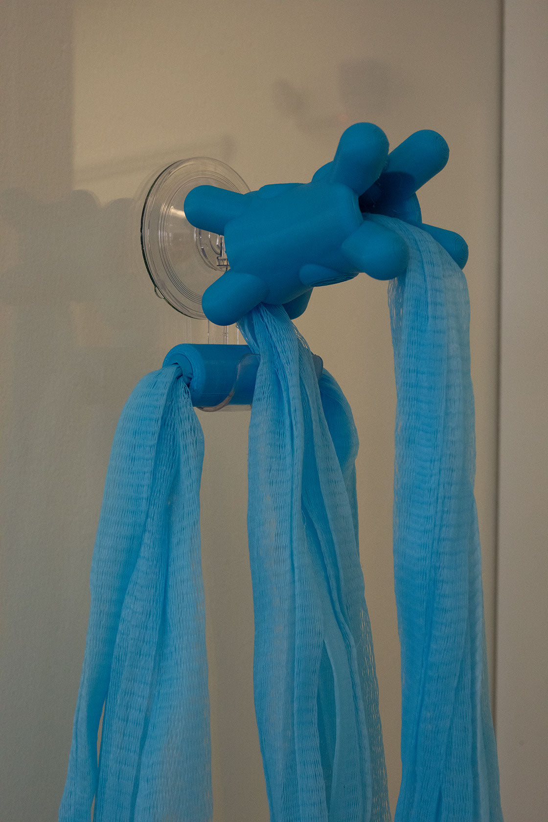 Blue Fähloo mesh and handles hanging from the clear Fähloo Shower Valet on a shower wall